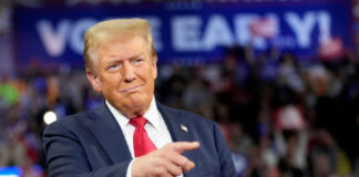 A man in a suit gestures while speaking at a political rally with a cheering crowd in the background