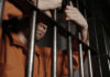 A male prisoner in an orange jumpsuit holding onto the bars of a jail cell