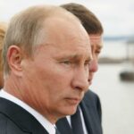 Close-up of a serious-looking man in a suit at an outdoor event near water