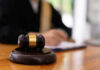 A gavel on a wooden table with a blurred figure of a person in professional attire in the background