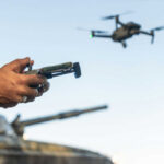 Person controlling a drone with a remote while a tank is in the background