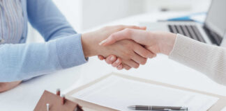 Two people shaking hands over a real estate contract with a model house in the foreground