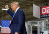 A man in a suit raises his fist while standing in a manufacturing facility with an American flag in the background
