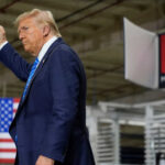 A man in a suit raises his fist while standing in a manufacturing facility with an American flag in the background