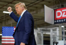 A man in a suit raises his fist while standing in a manufacturing facility with an American flag in the background