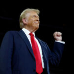 A man in a suit with a red tie raising his fist confidently during a speech