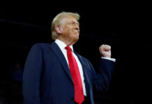 A man in a suit with a red tie raising his fist confidently during a speech