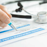 A hand holding a pen writing on a health insurance claim form with a stethoscope in the background