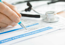 A hand holding a pen writing on a health insurance claim form with a stethoscope in the background