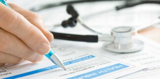 A hand holding a pen writing on a health insurance claim form with a stethoscope in the background
