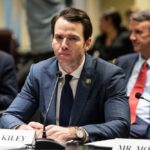 A man in a suit sitting at a table during a government hearing
