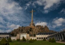 A large cross atop a rocky hill with a building in the foreground