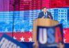 Astonishing Bid: Billionaire Governor Aims for Historic Third Term A politician delivering a speech at a podium with a backdrop of the USA flag