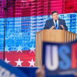 A politician delivering a speech at a podium with a backdrop of the USA flag