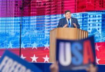 A politician delivering a speech at a podium with a backdrop of the USA flag