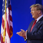 A man in a suit standing beside an American flag during a public speaking event