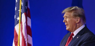 A man in a suit standing beside an American flag during a public speaking event