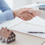 Two people shaking hands over a real estate contract with a model house in the foreground