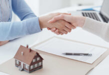 Two people shaking hands over a real estate contract with a model house in the foreground
