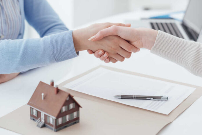 Two people shaking hands over a real estate contract with a model house in the foreground