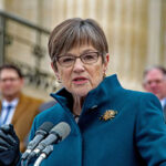 A woman in a teal coat speaking at a podium with microphones