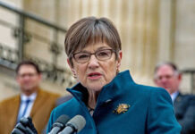 A woman in a teal coat speaking at a podium with microphones