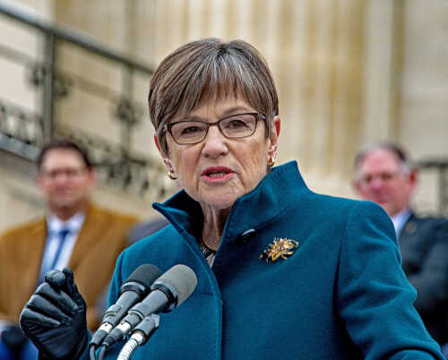 A woman in a teal coat speaking at a podium with microphones