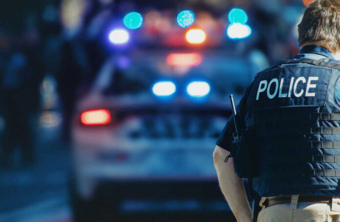 Police officer in a tactical vest standing in front of a police car with flashing lights