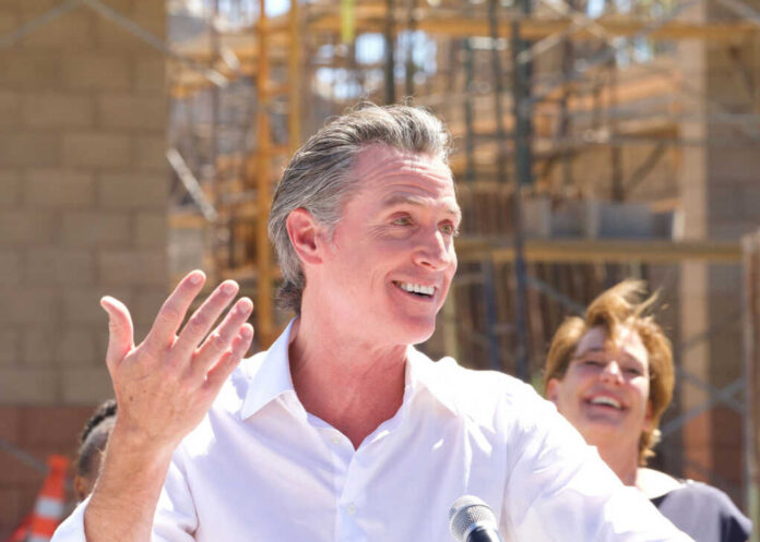 A man speaking at a construction site with a microphone, smiling, while a woman in the background laughs.