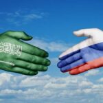 A green hand representing Saudi Arabia and a white hand representing Russia reaching out for a handshake against a blue sky