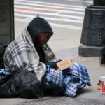 A homeless man sitting on the street wrapped in a blanket with a sign asking for help