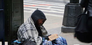 A homeless man sitting on the street wrapped in a blanket with a sign asking for help