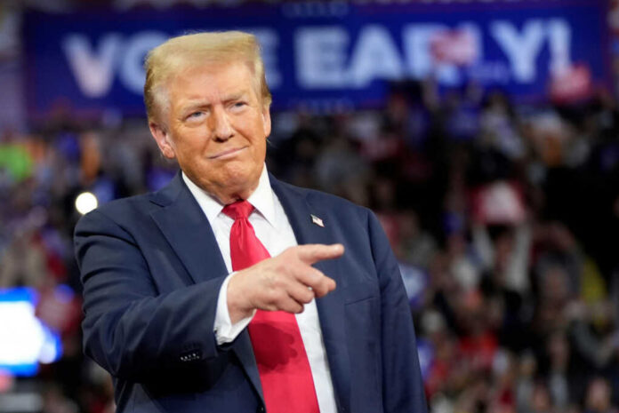 A man in a suit gestures while speaking at a political rally with a cheering crowd in the background
