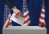 A hand placing a ballot into a ballot box with American flags in the background