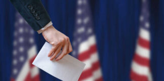 A hand placing a ballot into a ballot box with American flags in the background