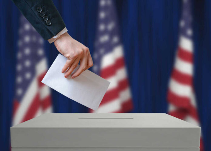 A hand placing a ballot into a ballot box with American flags in the background