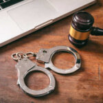 Handcuffs and a gavel placed next to a laptop on a wooden surface