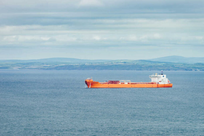 shutterstock_1695593065 An orange cargo ship sailing in the ocean with a coastal landscape in the background