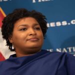 Abrams’ Political Future at Risk – Subpoena Drama! A political figure speaking at a podium with a blue backdrop