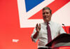 Market Panic: Speech Triggers Economic Jitters A man in a white shirt and tie gestures while speaking at a podium with a British flag backdrop