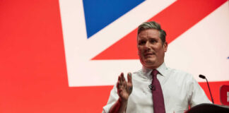 A man in a white shirt and tie gestures while speaking at a podium with a British flag backdrop