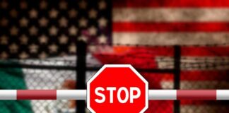 Stop sign on barrier with blurred U.S. and Mexico flags behind fence