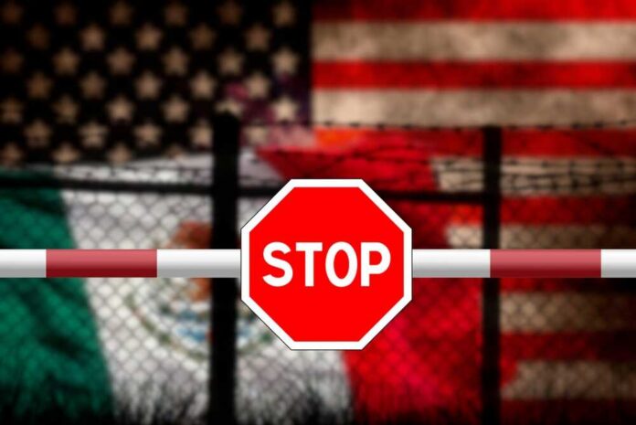 A,Fence,And,Barrier,With,A,Stop,Sign,At,The Stop sign on barrier with blurred U.S. and Mexico flags behind fence