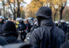 A crowd of masked individuals facing law enforcement in an autumn setting