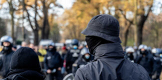 A crowd of masked individuals facing law enforcement in an autumn setting