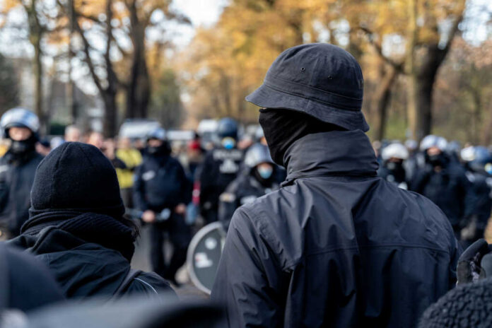shutterstock_1879968487 A crowd of masked individuals facing law enforcement in an autumn setting