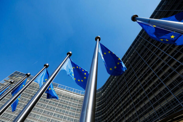 shutterstock_2317670971 European Union flags flying in front of a modern building against a clear blue sky