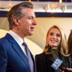 DOJ Firestorm Engulfs Newsom Man in suit speaking at a podium with American and California flags