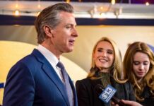 DOJ Firestorm Engulfs Newsom Man in suit speaking at a podium with American and California flags