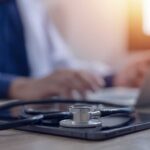 Berlin Doctor Convicted Of Fifteen Murders A stethoscope resting on a tablet with a blurred medical professional in the background typing on a laptop